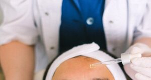 Top view skilled female cosmetologist in uniform and gloves applying essential oil on young woman face skin while working in contemporary spa center